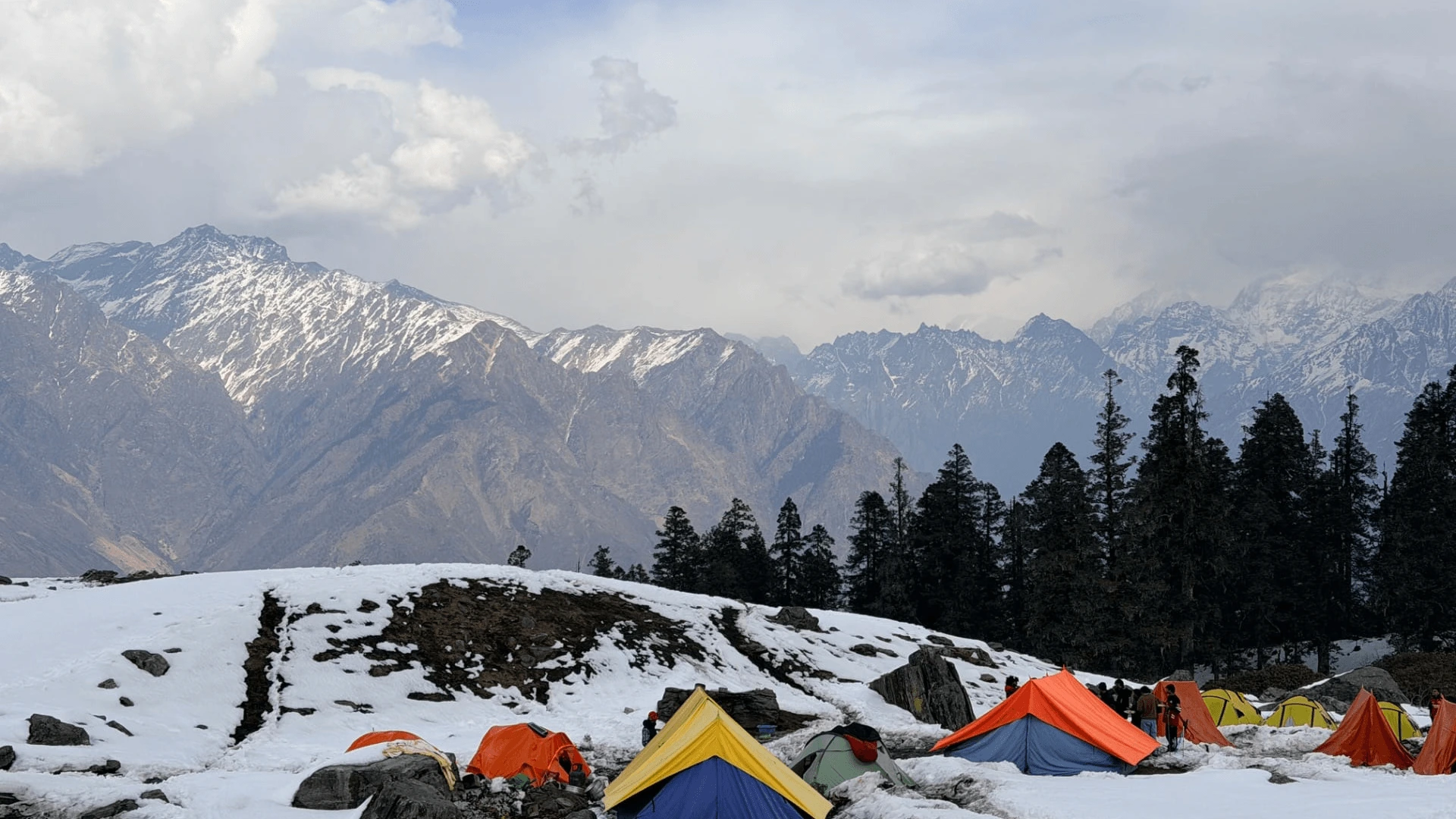 Colorful trekking tents at a snow-covered Himalayan campsite surrounded by pine forests and majestic mountain peaks.